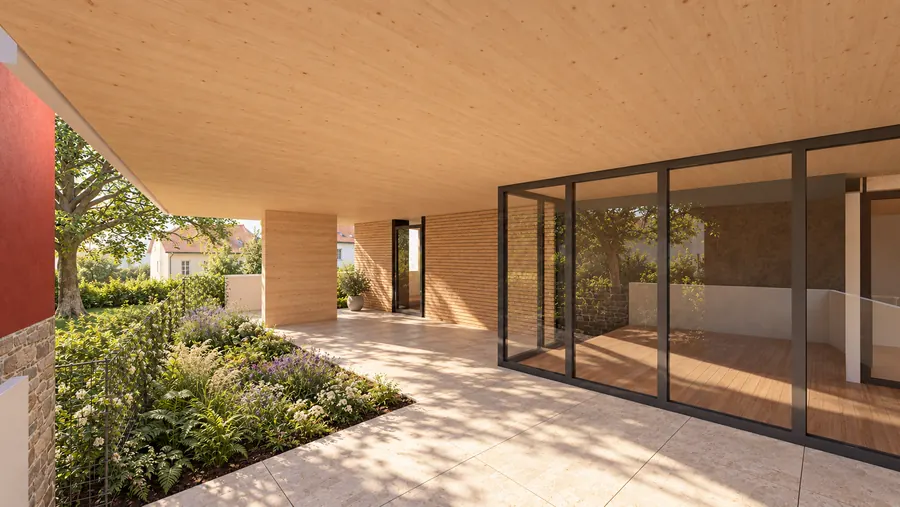 Eine überdeckte Terrasse unter der Decke aus Brettsperrholz: links eine rot verputzte Wand, ein Baum und ein Beet mit blühenden Stauden hinter einem Gitter, rechts die Glaswand zu einem Raum mit Dielenboden; dahinter die Hecke und das Nachbarhaus mit Ziegeldach.